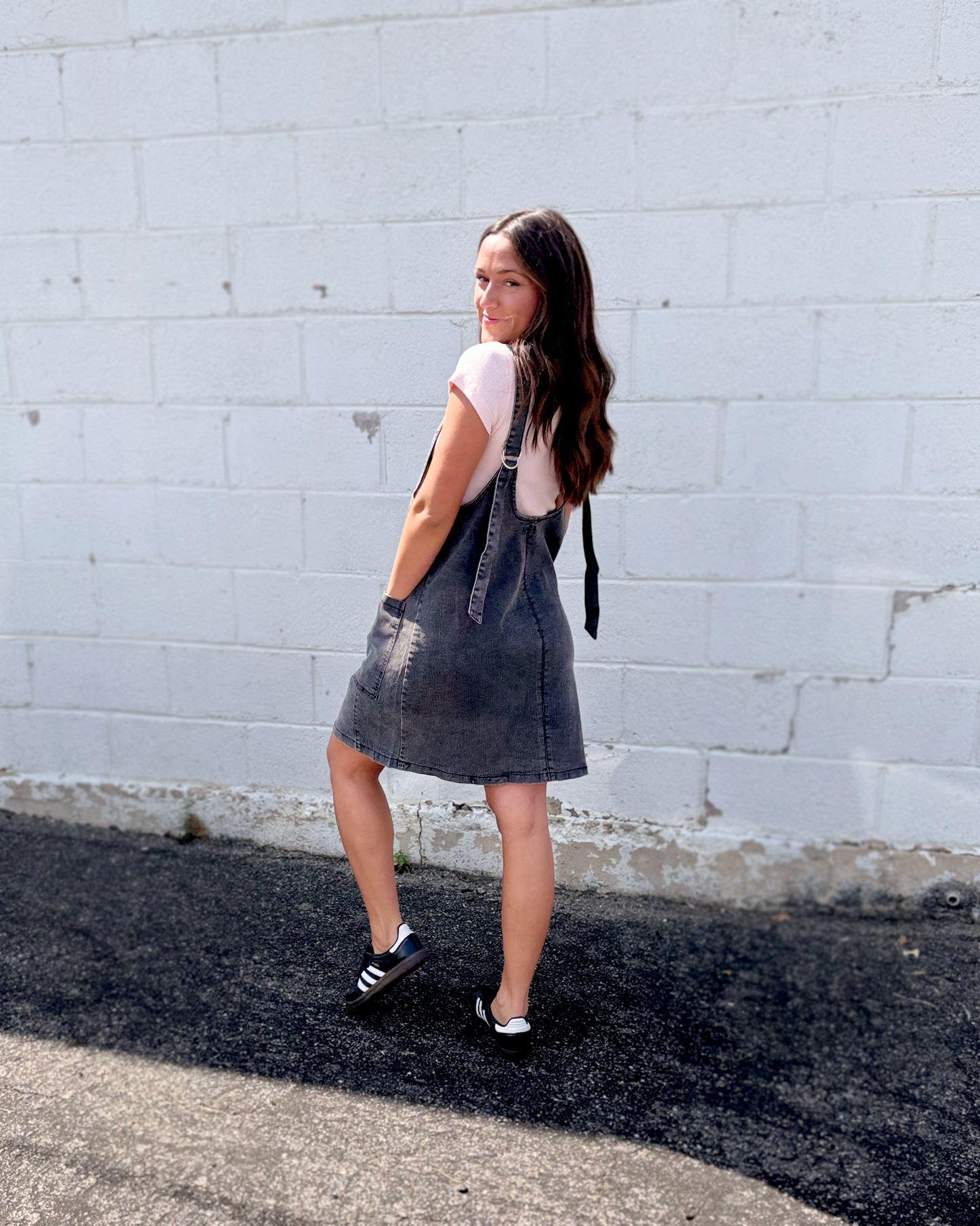 A woman stands outdoors against a white painted brick wall, wearing a dark gray denim overall dress over a light pink short-sleeve top, with black and white sneakers.